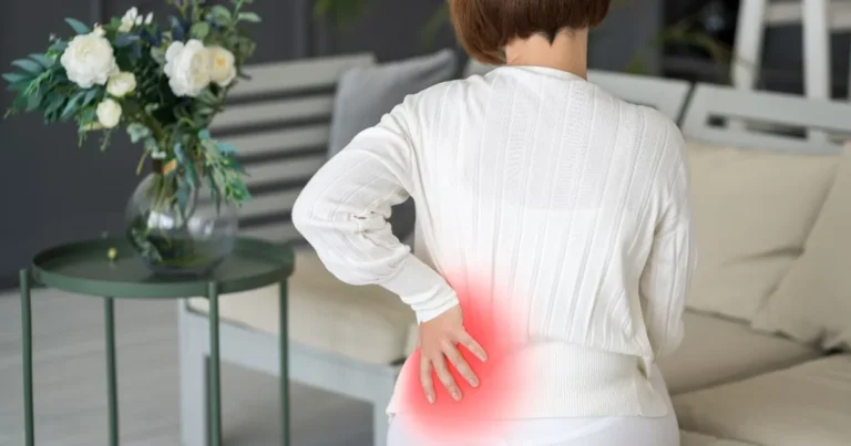 Woman in a white sweater sitting on a couch, holding her lower back in pain, with a red highlight indicating discomfort,when does tailbone pain start in pregnancy