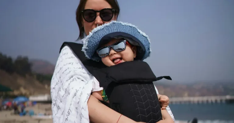 Baby wearing sunglasses and a blue hat in a baby carrier on the beach, associated with names that mean death ,baby carrier seat