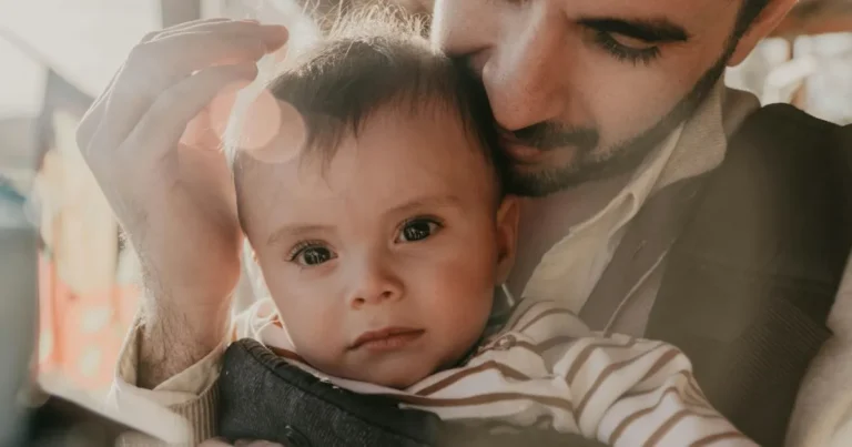 A tender moment between a father and his baby, with the father gently cradling the child in his arms. The sunlight creates a soft lens flare, highlighting the baby's curious expression and the father's loving gaze. ,baby carrier for dad