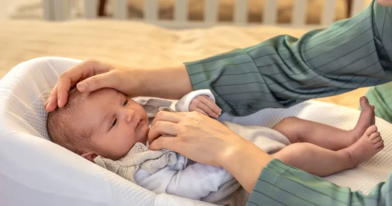 Newborn baby lying in a soft white baby lounger with a parent's hand gently placed on their forehead, suggesting care or checking for fever, newborn won't sleep in bassinet