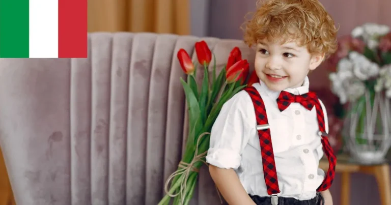 A young boy with curly blond hair, dressed in a white shirt with red plaid suspenders and a matching bow tie, sits on a plush gray chair. He smiles brightly while holding a bunch of red tulips behind his back. An Italian flag is visible in the upper-left corner of the image., italian boy names
