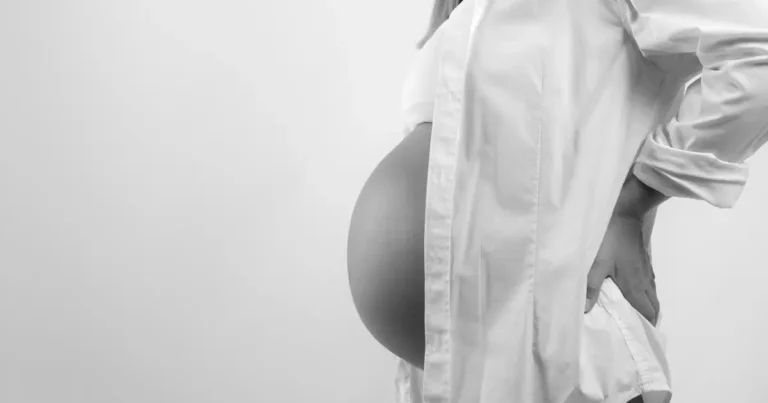 Silhouette of a pregnant woman standing in front of a window, cradling her belly with both hands, Pregnancy Pain in the Coccyx