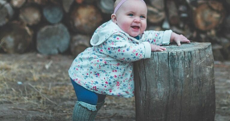 Smiling baby girl in floral hoodie and blue tights standing by a wooden stump in an outdoor forest setting., names starting with v