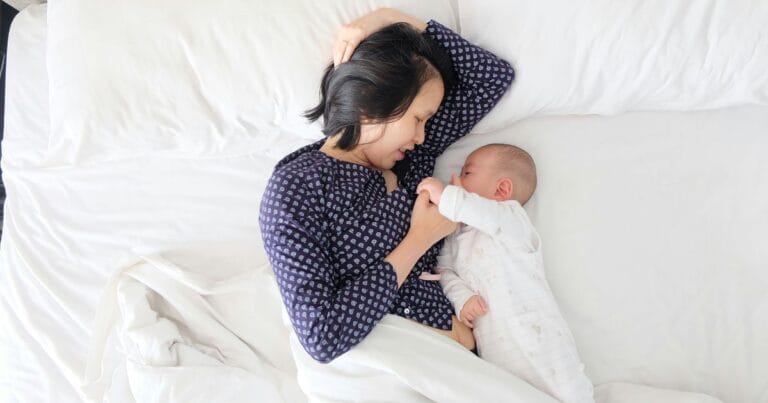 Mother breastfeeding baby in bed under white blanket, How to Get an Overtired Baby to Sleep
