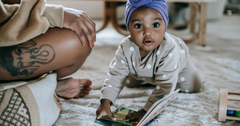 urious baby girl in polka dot outfit and purple turban crawling on carpet with a picture book, mother beside her with visible tattoo ,Mixed Race Names