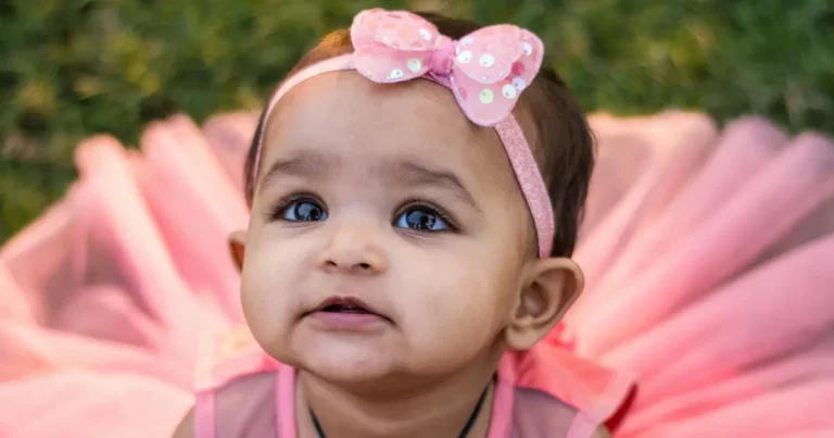 A baby girl with big expressive eyes wears a pink bow headband and a pink outfit while sitting on the grass, looking upward with curiosity. ,what is a good cherokee baby names