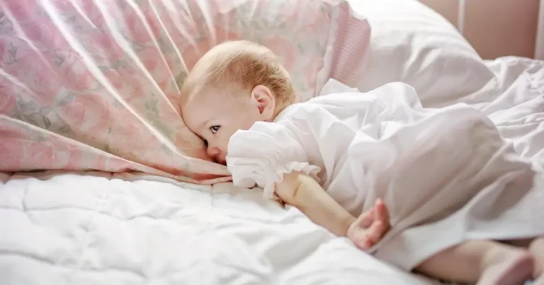 Baby in white dress lying on a floral pillow, looking wide-eyed and relaxed, when can babies sleep on their stomach