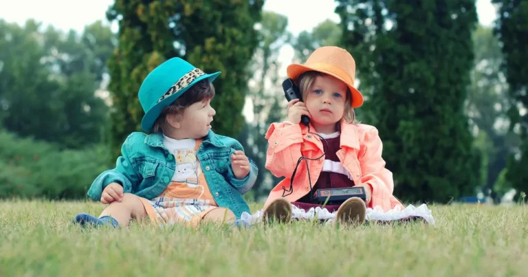Two toddlers sitting on grass, wearing colorful hats and jackets, one child holding a vintage telephone receiver to their ear while the other looks on, Italian names