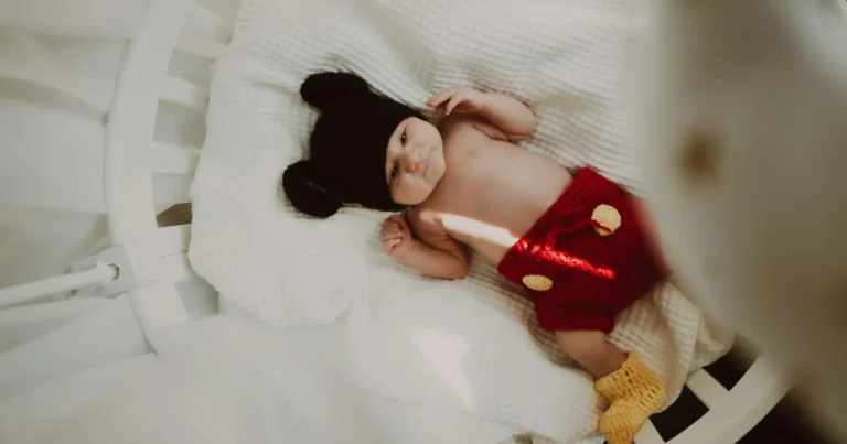 Baby lying in crib dressed in red knitted shorts, yellow booties, and black hat with mouse ears, looking up calmly, how to get newborn to sleep in crib