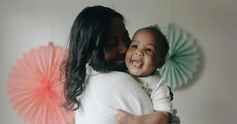 Black baby boy smiling while being held by his mother, wearing a white outfit with colorful decorations in the background , black boy names.