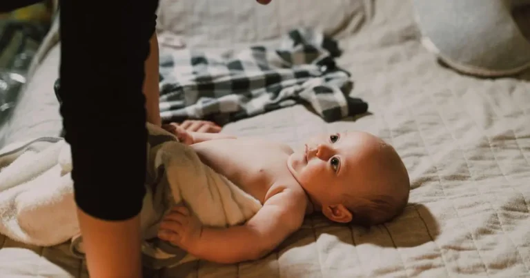 girl names that start with k. A baby girl with a pink bow headband lying on a white blanket, smiling gently at the camera.