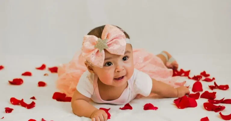 Baby in a sparkly dress with a large bow headband, lying among red rose petals – names that start with rose