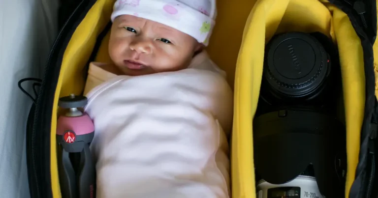 A swaddled newborn baby lies inside a padded camera bag next to professional photography gear, including a Canon lens and a mini tripod, blending baby care with photography essentials.