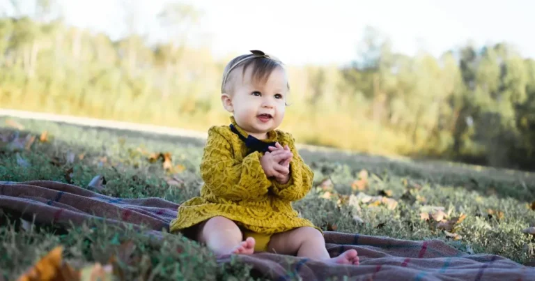 A smiling baby girl in a mustard yellow dress sits on a blanket in a grassy fieldj girl names