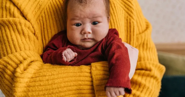 Newborn baby with scrunched face in red outfit held in yellow blanket, demonstrating signs of newborn scrunch reflex