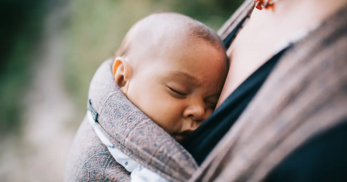a mother her carrying a sleeping daughter snuggled inside a soft linen baby wrap,