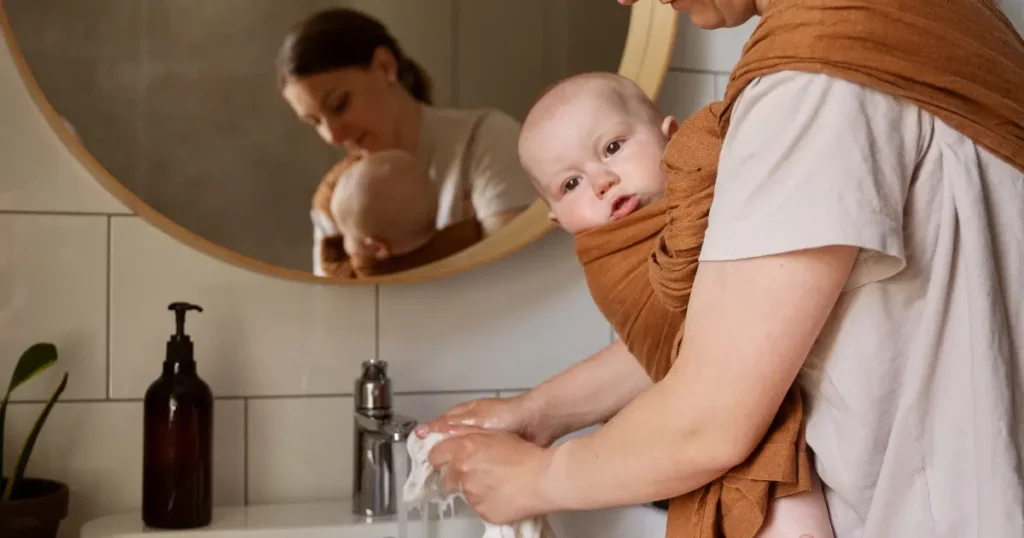 A medium shot shows a woman doing household chores while keeping her baby close and secure in a linen baby wrap.