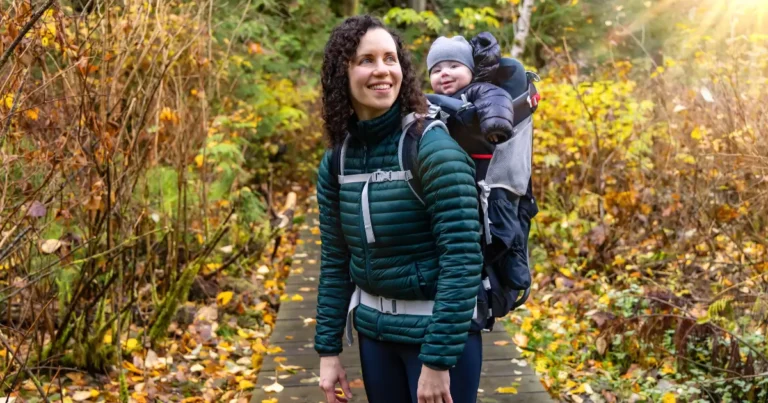 Mother hiking on a forest trail during fall, smiling with her Hiking Backpack for Toddlers. Both are warmly dressed for cool weather.