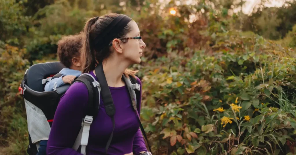 Mother hiking through a lush trail at sunset with her toddler riding in a hiking backpack carrier. She wears a purple top and looks ahead thoughtfully.