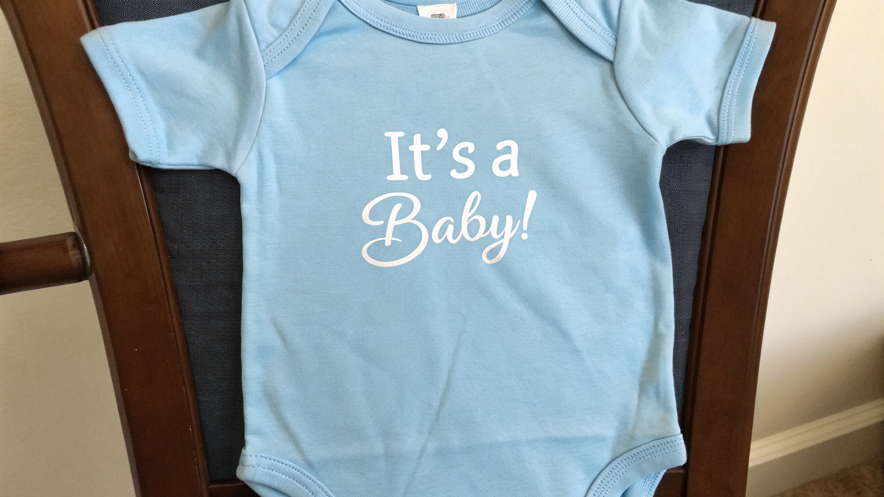 Blue pregnancy announcement onesie with the text It’s a Baby! displayed on a chair.