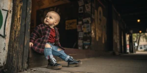 A baby dressed in a Grunge Names outfit with an oversized flannel, ripped jeans, and tiny Doc Martens boots, sitting in front of a dark 90s-style textured background with graffiti vibes.